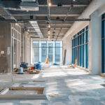Open office space under renovation with exposed ceiling, scattered materials, and blue-framed windows bringing in daylight