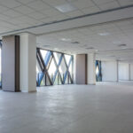 Empty open-plan office space with white columns and floor-to-ceiling diagonal-window walls looking out on greenery