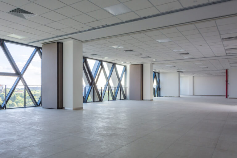 Empty open-plan office space with white columns and floor-to-ceiling diagonal-window walls looking out on greenery