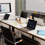 Long conference table with three open laptops, papers, charts, and coffee cups in a bright meeting room with a large monitor in the background