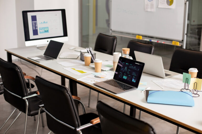 Long conference table with three open laptops, papers, charts, and coffee cups in a bright meeting room with a large monitor in the background