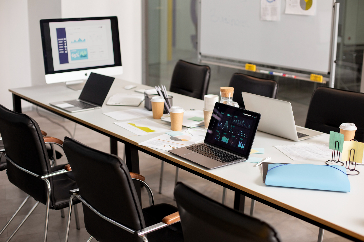 Long conference table with three open laptops, papers, charts, and coffee cups in a bright meeting room with a large monitor in the background